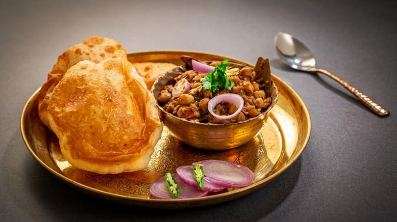 A golden plate holds chole bhature, a bowl of chickpeas, and sliced onions with green chilies.