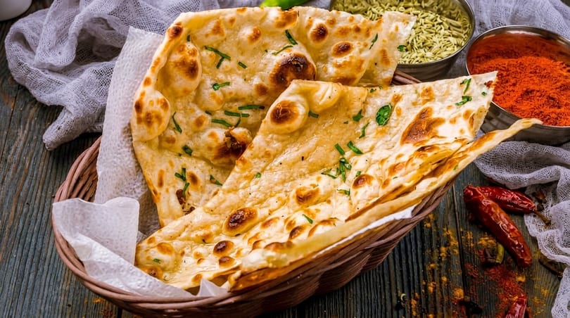 Basket of golden naan bread with herbs, alongside bowls of spices and dried chilies.